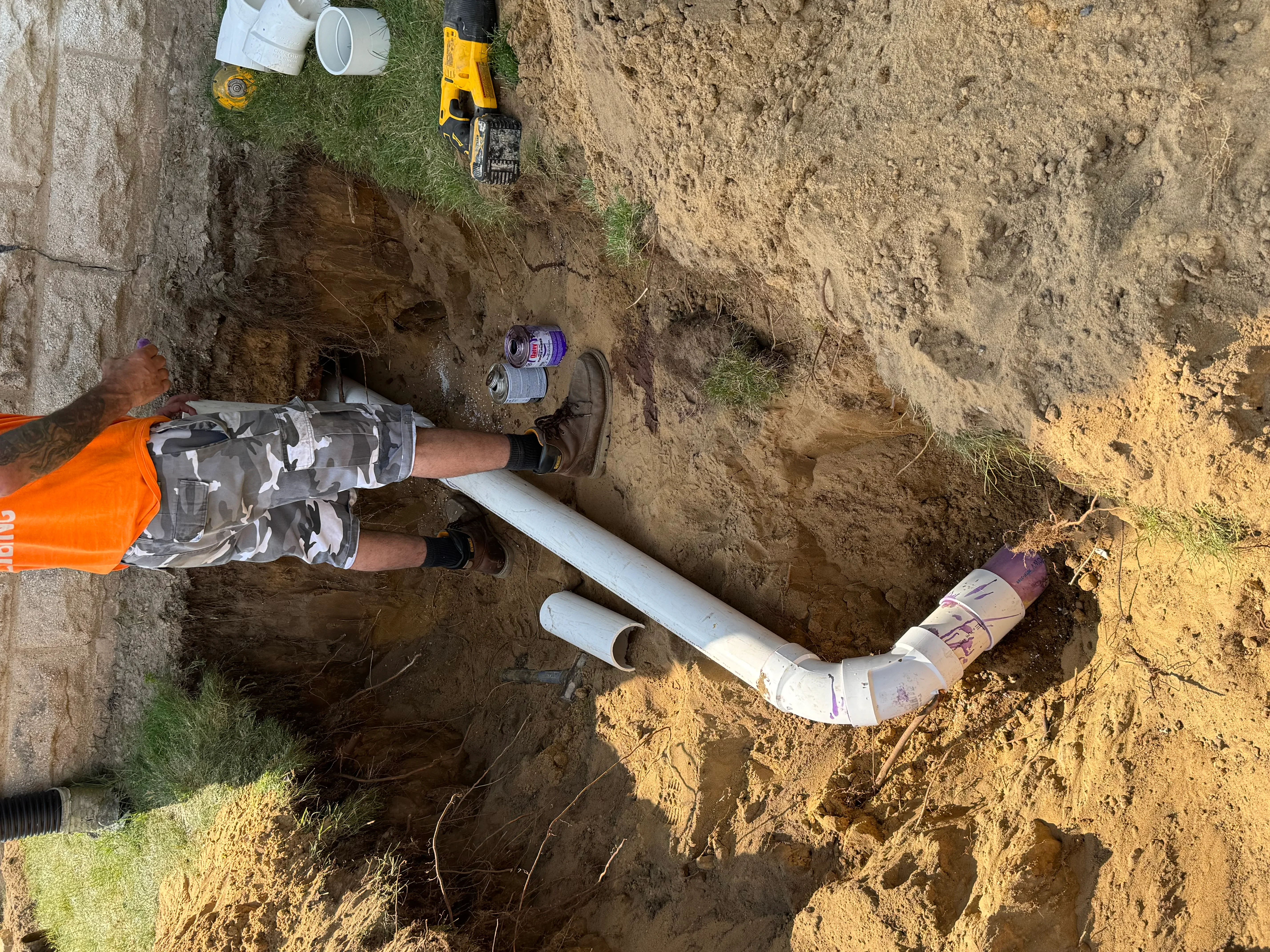 Worker installing white PVC drainage pipe with elbow fitting in excavated trench