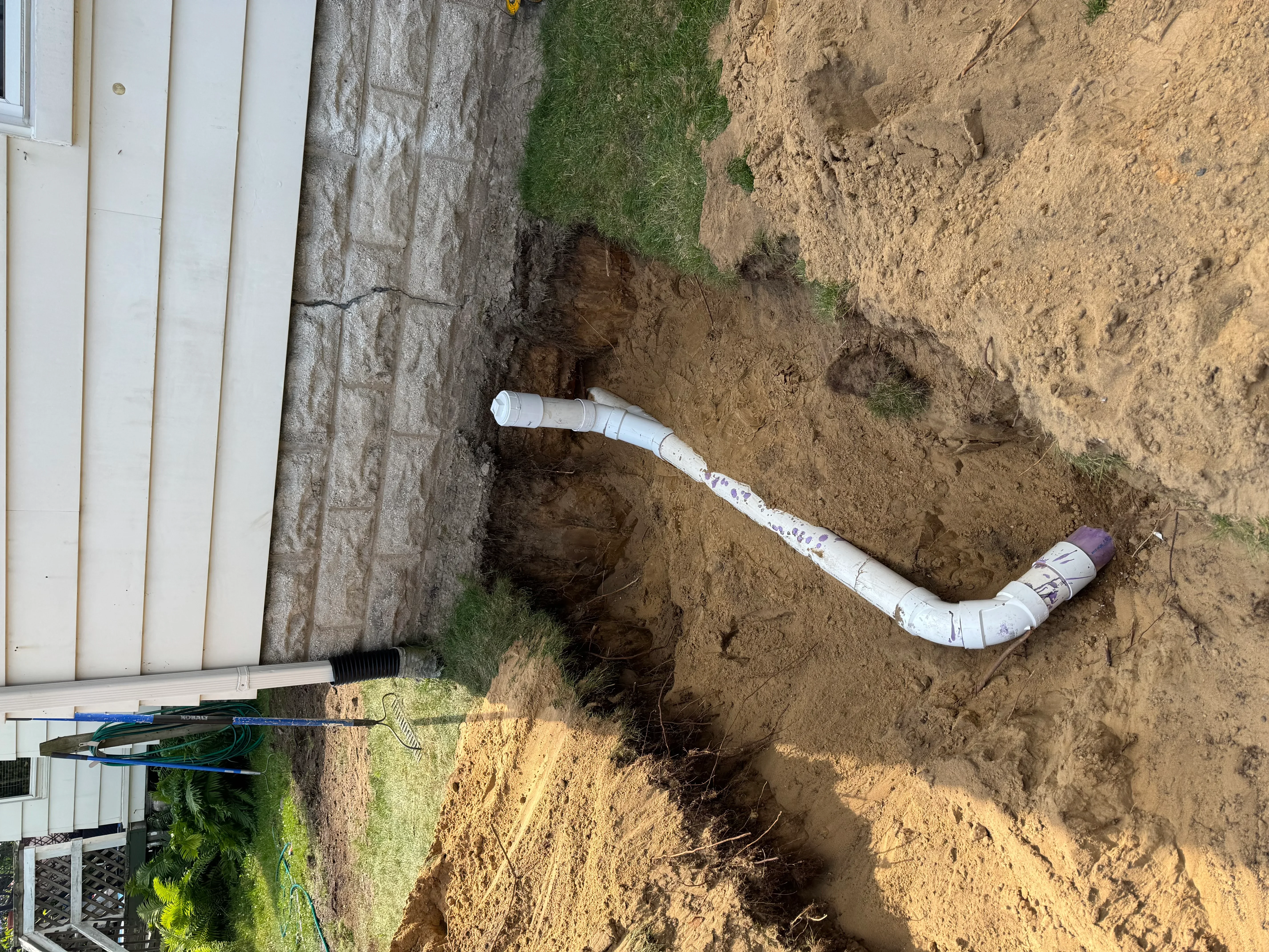 White PVC drainage pipe with purple coupling installed in excavated trench next to foundation