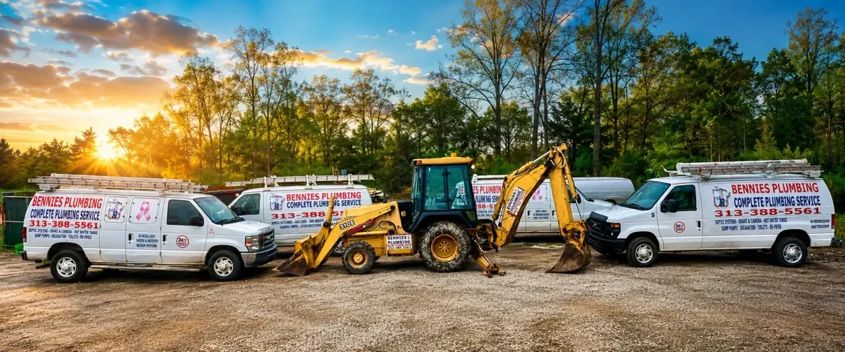 White plumbing service vans and yellow backhoe excavator lined up in equipment yard at sunset