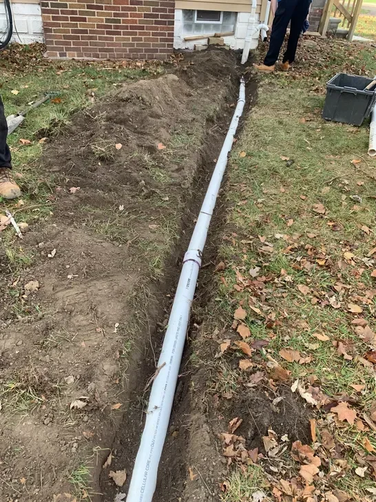 Gray PVC drain pipe installed in excavated trench along brick home foundation